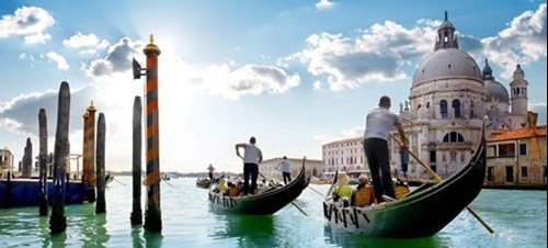 Two gondolas with tourists sail toward the Basilica of Santa Maria della Salute on a sunny day.
