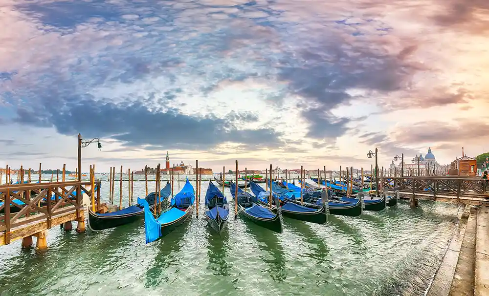 Rows of blue gondolas moored along Riva degli Schiavoni, with San Giorgio Island in the distance and a cloudy dawn sky