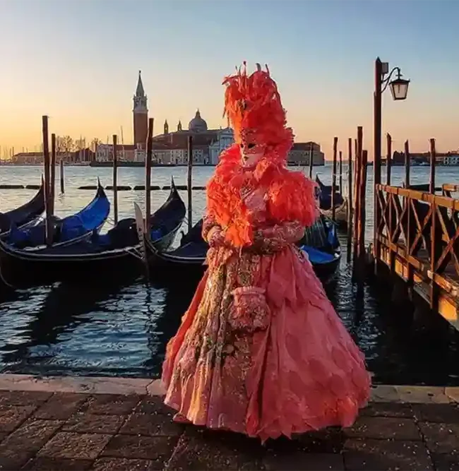 A costumed figure in pink stands on the Venetian waterfront, with moored gondolas and San Giorgio Maggiore at sunset.