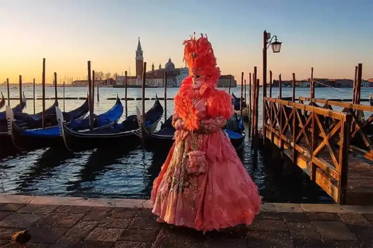 A costumed figure in pink stands on the Venetian waterfront, with moored gondolas and San Giorgio Maggiore at sunset.