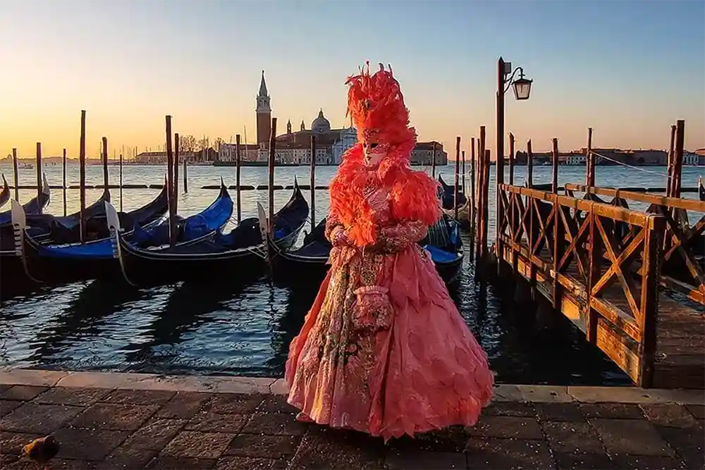 Persona mascherata in elegante abito rosa e piume, fotografata al tramonto con le gondole e San Giorgio sullo sfondo